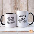 Two customizable white ceramic mugs with black handles and interiors, displayed side-by-side on a rustic white wooden surface with a weathered wood background. The mugs feature placeholder black text reading 'ADD YOUR CUSTOM TEXT HERE'.