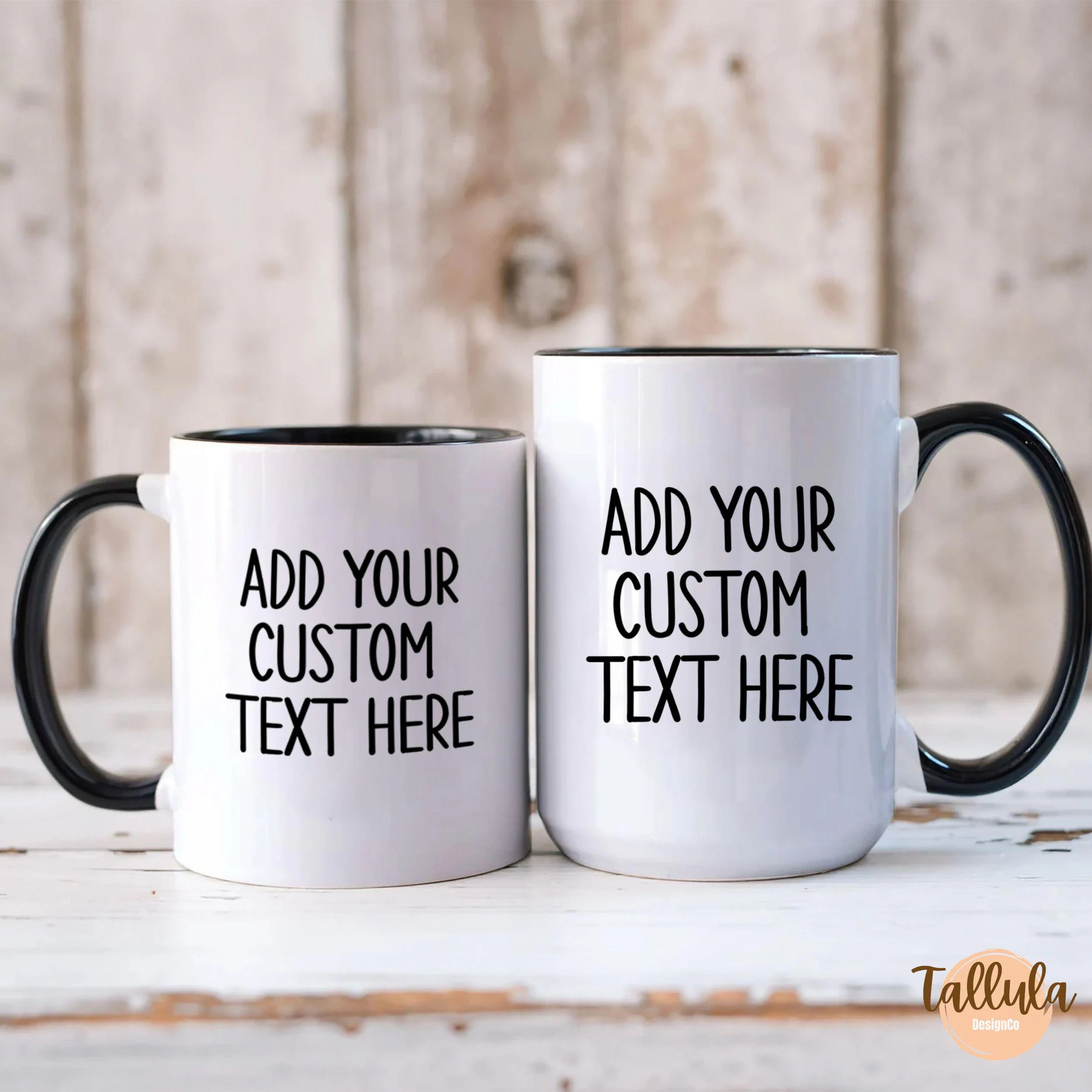 Two customizable white ceramic mugs with black handles and interiors, displayed side-by-side on a rustic white wooden surface with a weathered wood background. The mugs feature placeholder black text reading 'ADD YOUR CUSTOM TEXT HERE'.