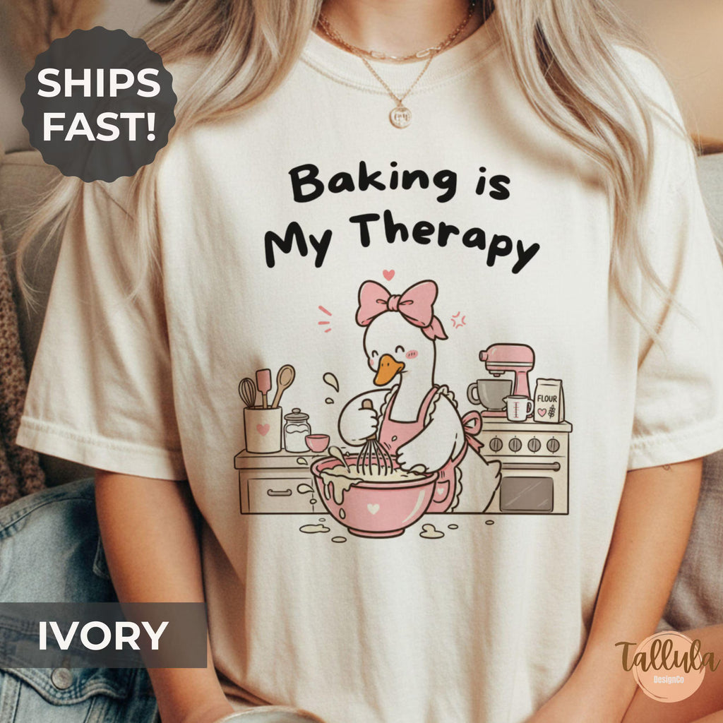 A graphic T-shirt with the text 'Baking is My Therapy' and a cartoon goose baking illustration with messy kitchen bench
