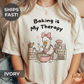 A graphic T-shirt with the text 'Baking is My Therapy' and a cartoon goose baking illustration with messy kitchen bench