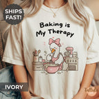 A graphic T-shirt with the text 'Baking is My Therapy' and a cartoon goose baking illustration with messy kitchen bench