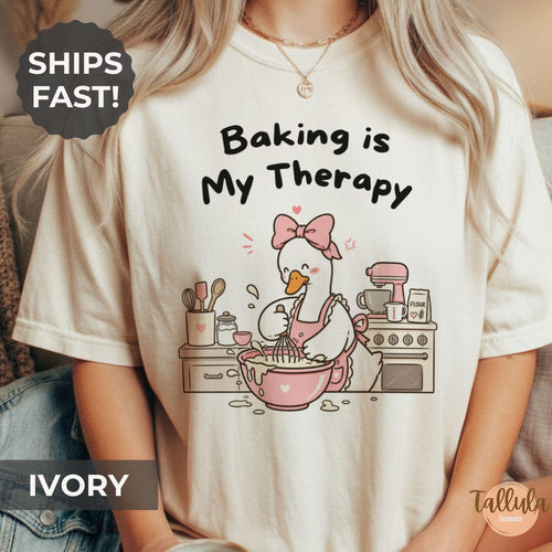 A graphic T-shirt with the text 'Baking is My Therapy' and a cartoon goose baking illustration with messy kitchen bench