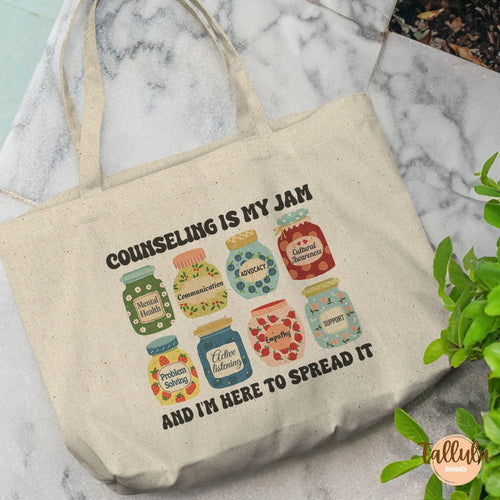 A natural canvas tote bag with printed text and illustrations of colorful jam jars, The text reads 'Counseling IS MY JAM' and 'AND I'M HERE TO SPREAD IT' with descriptive words related to therapist, school counseling, psychologist