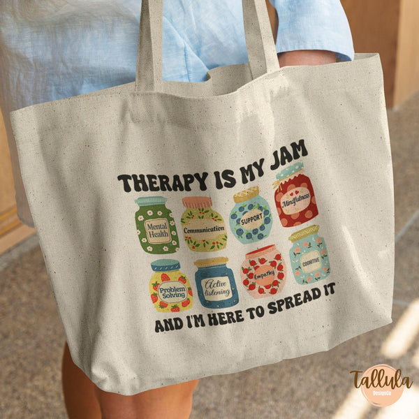 A natural canvas tote bag with a printed graphic of colorful jars and the text 'THERAPY IS MY JAM AND I'M HERE TO SPREAD IT'. Featuring the descriptive words related to therapy, counseling, mental health.
