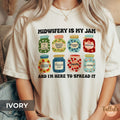 A comfort colors graphic t-shirt with the text 'MIDWIFERY IS MY JAM AND I'M HERE TO SPREAD IT' and eight colorful illustrated jam jars, featuring descriptive words on each jar related to midwife, doula, mother baby nurse, postpartum nurse, perinatal nurse
